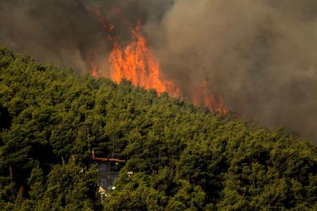 Las llamas y el denso humo de un incendio forestal elevándose sobre una casa en la zona de Porto Germeno&nbsp;